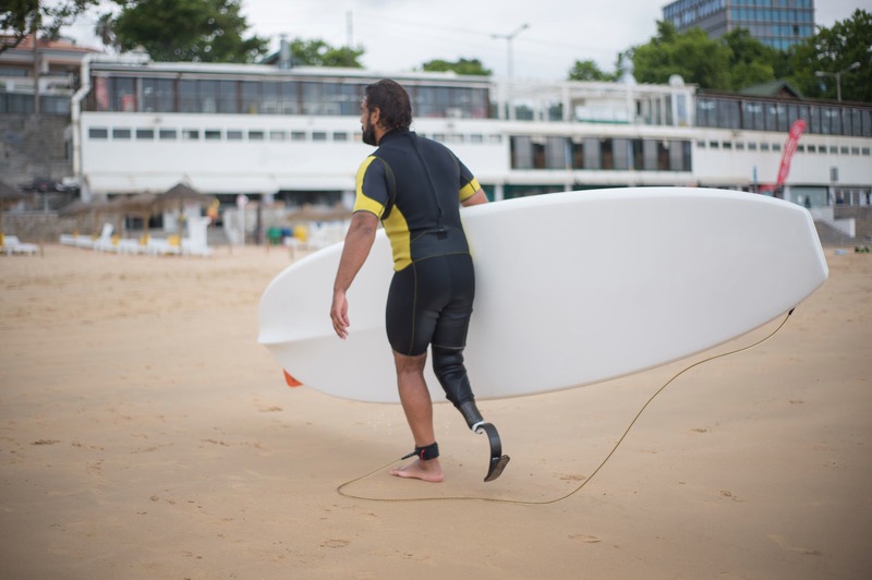 A man with a prosthetic leg carries a surfboard across the sand.