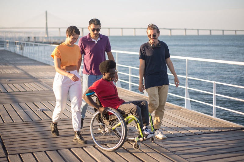 Four friends, one using a wheelchair, laugh together on a seaside boardwalk.