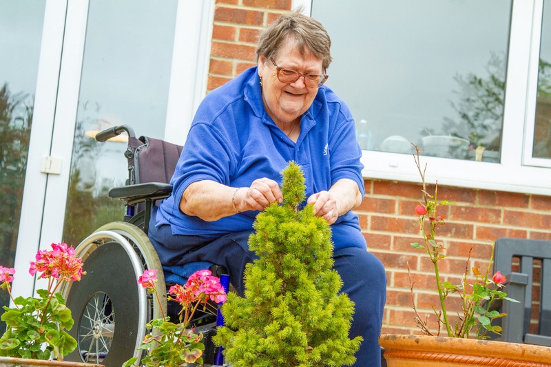 An older woman in a wheelchair laughs as she trims a shrub in her garden.