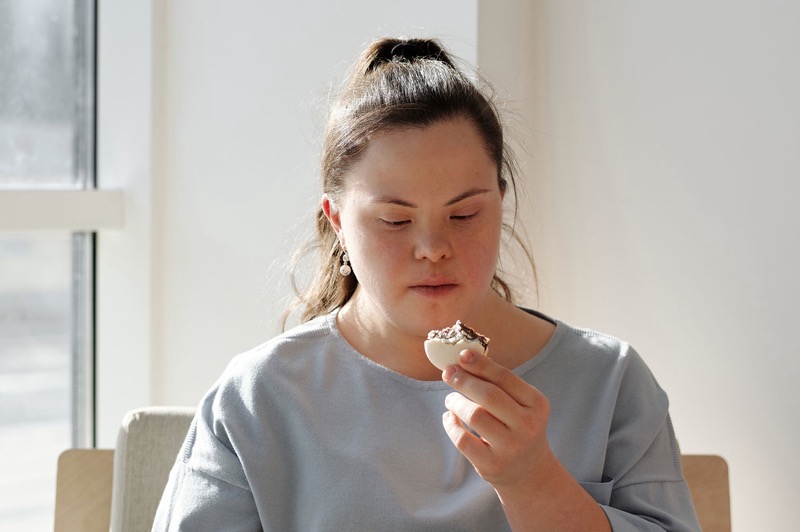 A young woman eats breakfast at a table by a sunny window.